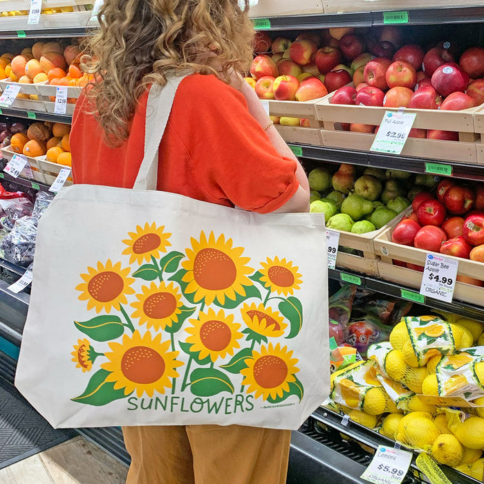 A woman shops for produce at the grocery store with The Neighborgoods Sunflower tote bag on her shoulder.