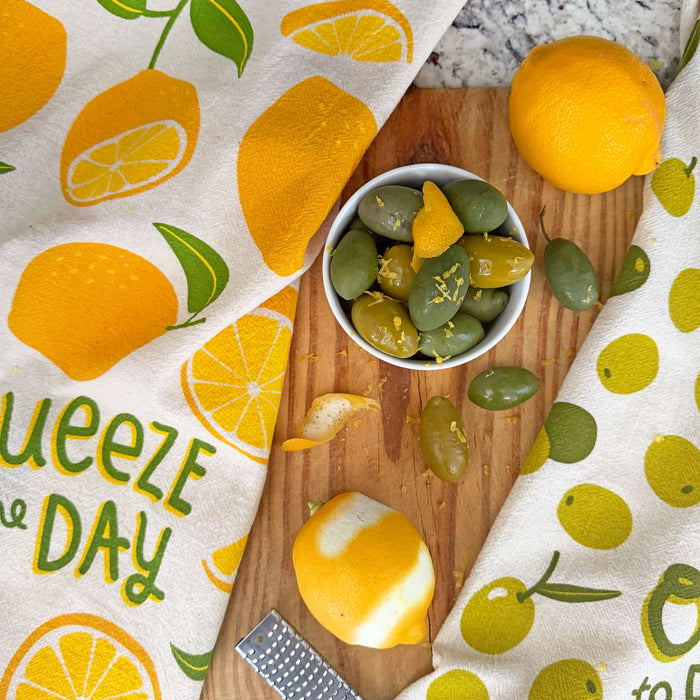 The Neighborgoods Lemon and Olive dish towels laid side by side on a kitchen countertop. A bowl of olives sprinkled with fresh lemon zest sits in between the dish towels.