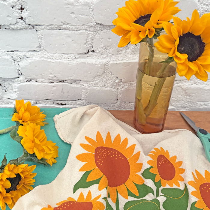 The Neighborgoods Sunflowers dish towel featuring a sunflower illustration, laid next to a vase of freshly cut sunflowers.