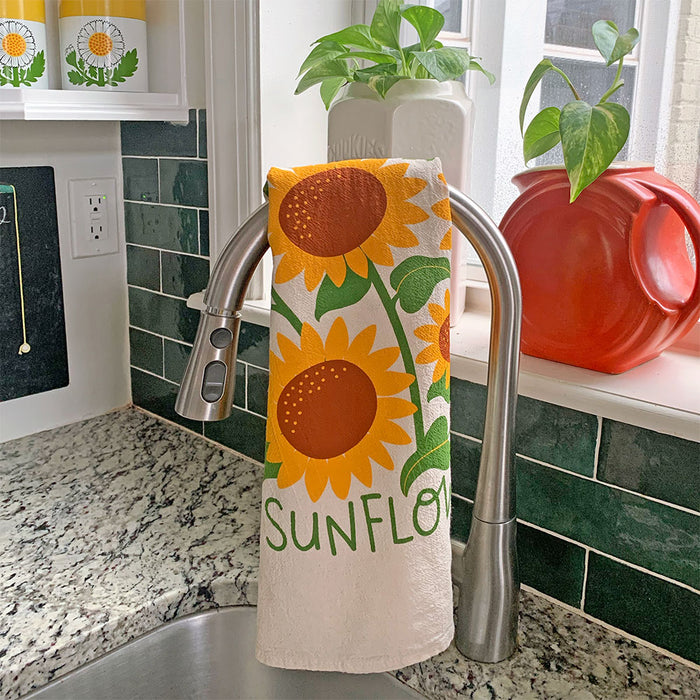 The Neighborgoods Sunflowers dish towel with a sunflower illustration hanging over a kitchen sink faucet.