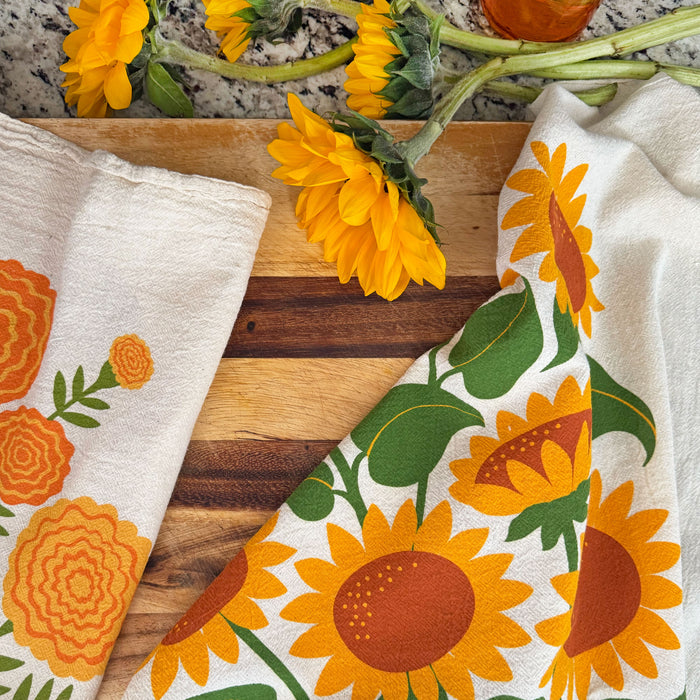 The Neighborgoods Sunflowers dish towel featuring a sunflower illustration, laid next to freshly cut sunflowers on a kitchen countertop.
