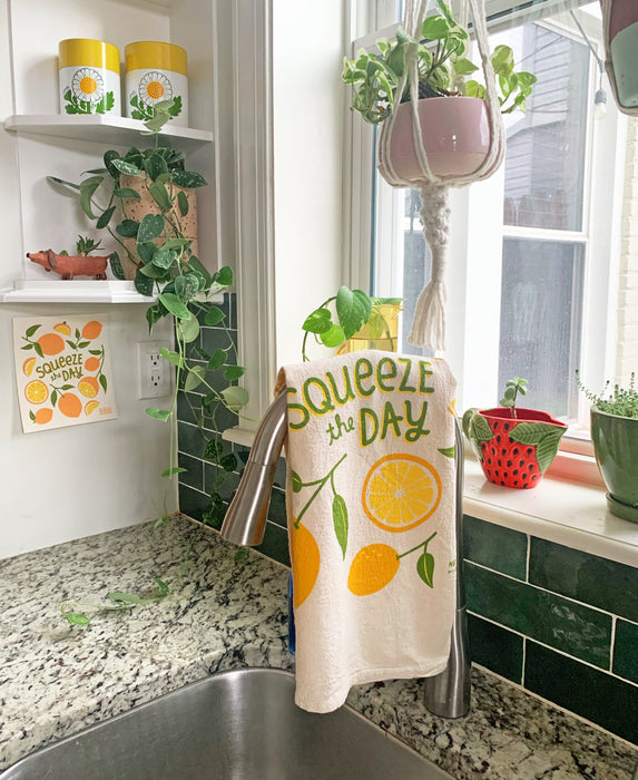 The Neighborgoods Lemon dish towel with a Squeeze the Day lemon food pun and lemon art illustration hanging over a kitchen sink faucet.
