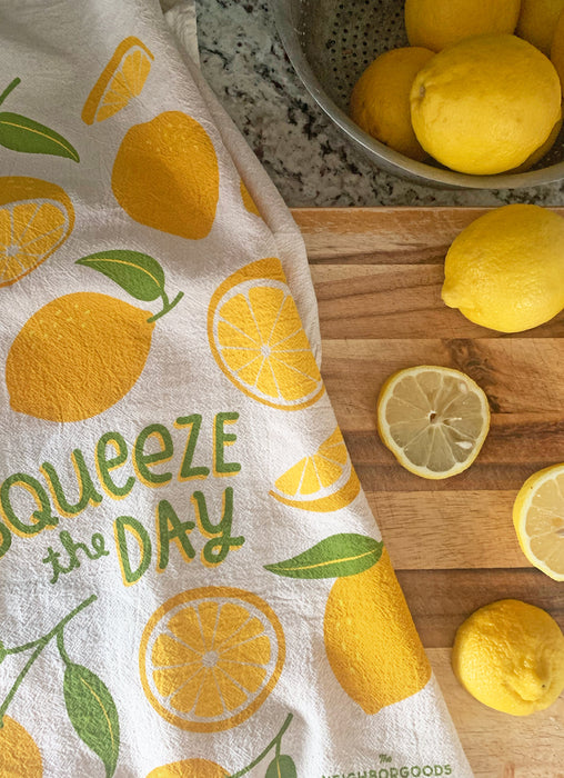 The Neighborgoods Lemon dish towel featuring a lemon illustration, laid next to freshly sliced lemons on a kitchen cutting board.