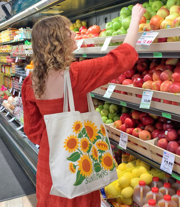 A woman shops for produce at the grocery store with The Neighborgoods Sunflower tote bag on her shoulder.