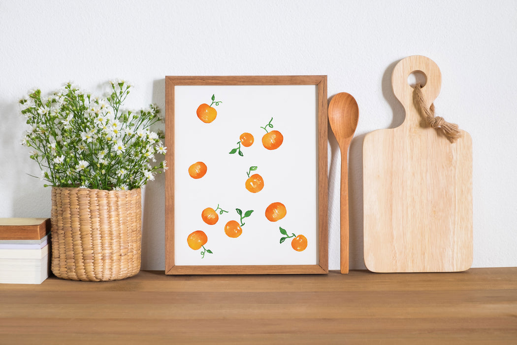 Watercolor clementines print in small size framed alongside a wooden spoon, cutting board, and flowering plant