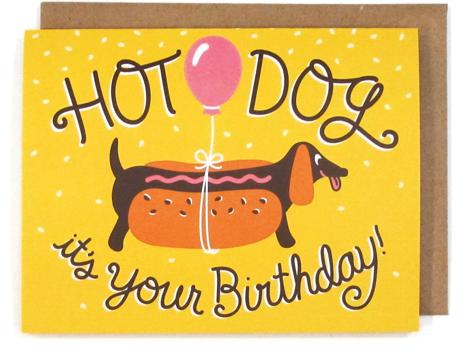 Yellow card that pictures a dachshund ina hot dog costume with a ballomn tied around its belly, featuring the phrase "Hot dog it's your birthday!," with a brown envelope
