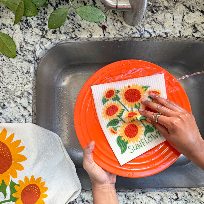 A woman washes a plate in the sink with a sunflower sponge cloth.