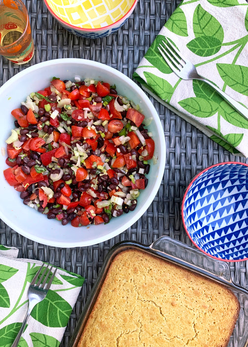 Two dishes on a table. One dish contains a black bean and tomato salad and another dish is a loaf of cornbread.