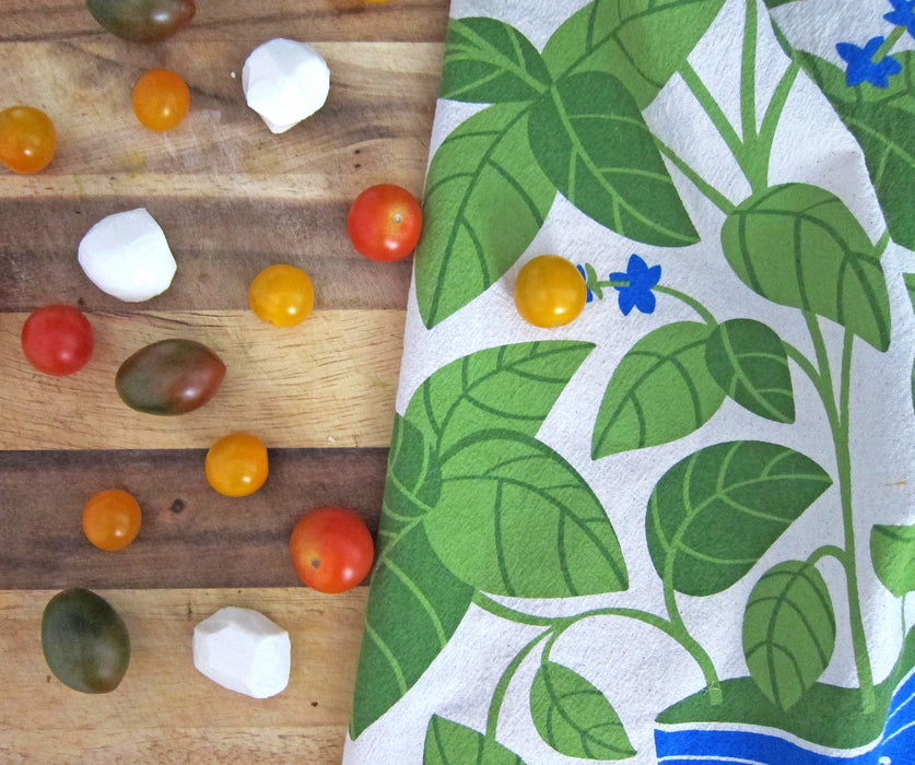 A basil dish towel draped over a cutting board with mozzarella balls and cherry tomatoes. 
